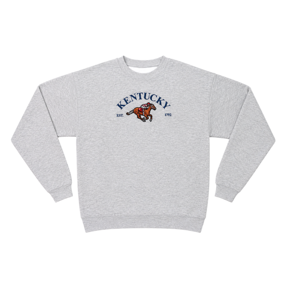 A light heather gray crewneck sweatshirt featuring a vintage-style graphic on the chest with the word 'KENTUCKY' in blue arched text, 'EST. 1992' in smaller blue text, and a brown and orange running bucking horse logo in the center.